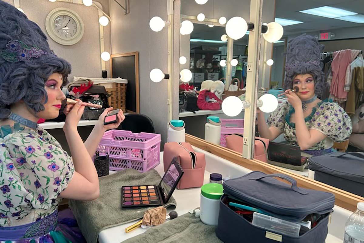 An actress in costume applies makeup to her face in front of a mirror.